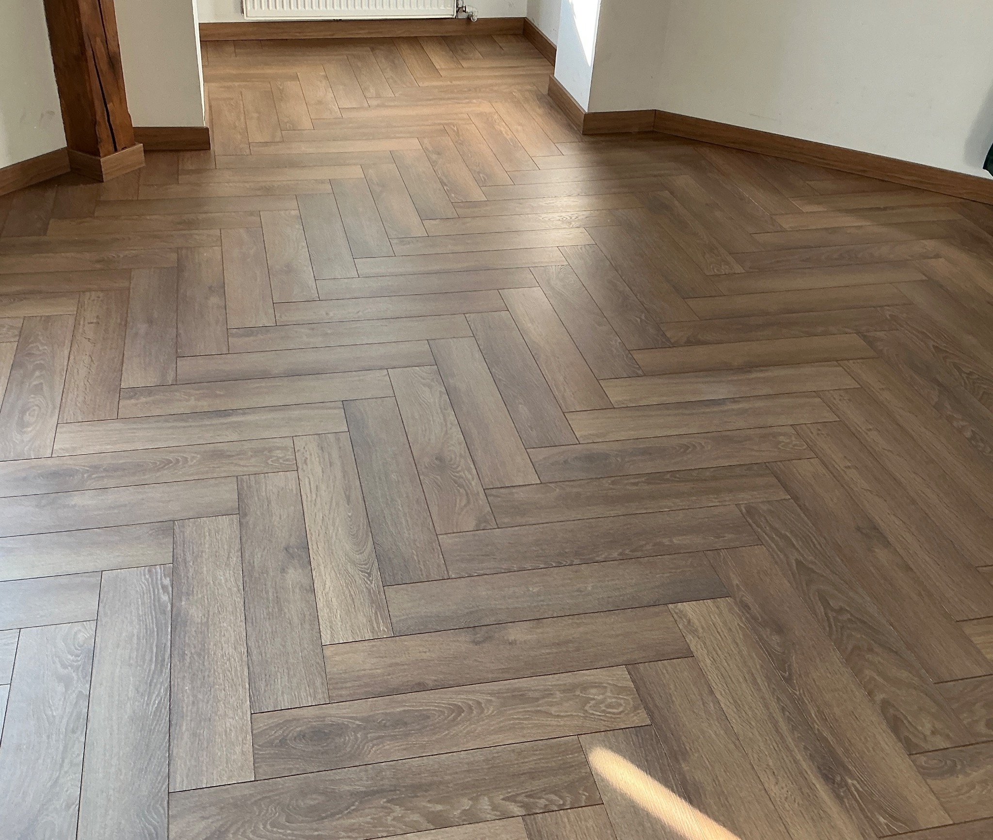 Ułożone panele podłogowe w jodełkę, z widocznymi listwami przypodłogowymi i fragmentem kaloryfera. Naturalne światło podkreśla wzór paneli.