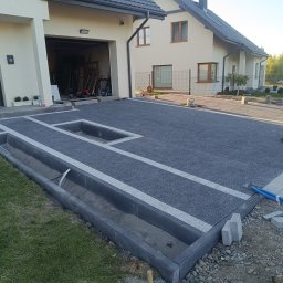 TerraFlo - Nowoczesny podjazd z kostki brukowej w trakcie układania, z widocznymi obrzeżami i miejscem na rośliny. W tle dom jednorodzinny i osoba w oddali.