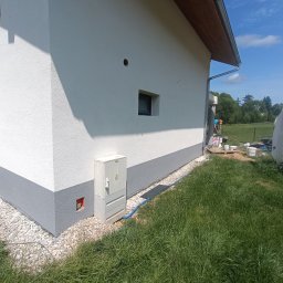 TerraFlo - Ocieplony budynek z pomalowaną elewacją na biało i szaro, widoczne elementy instalacji, trawnik i pracownik w tle wykonujący prace wykończeniowe przy budynku.