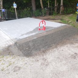 TerraFlo - Nowa, jasnoszara kostka brukowa na parkingu, z czerwonym blokadą na miejscu postojowym, otoczona szarym żwirem i zielenią w tle.