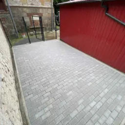 Świeżo ułożona, szara kostka brukowa na podwórku między budynkiem z cegły a czerwonym budynkiem gospodarczym. Widoczna brama z czarnej siatki.