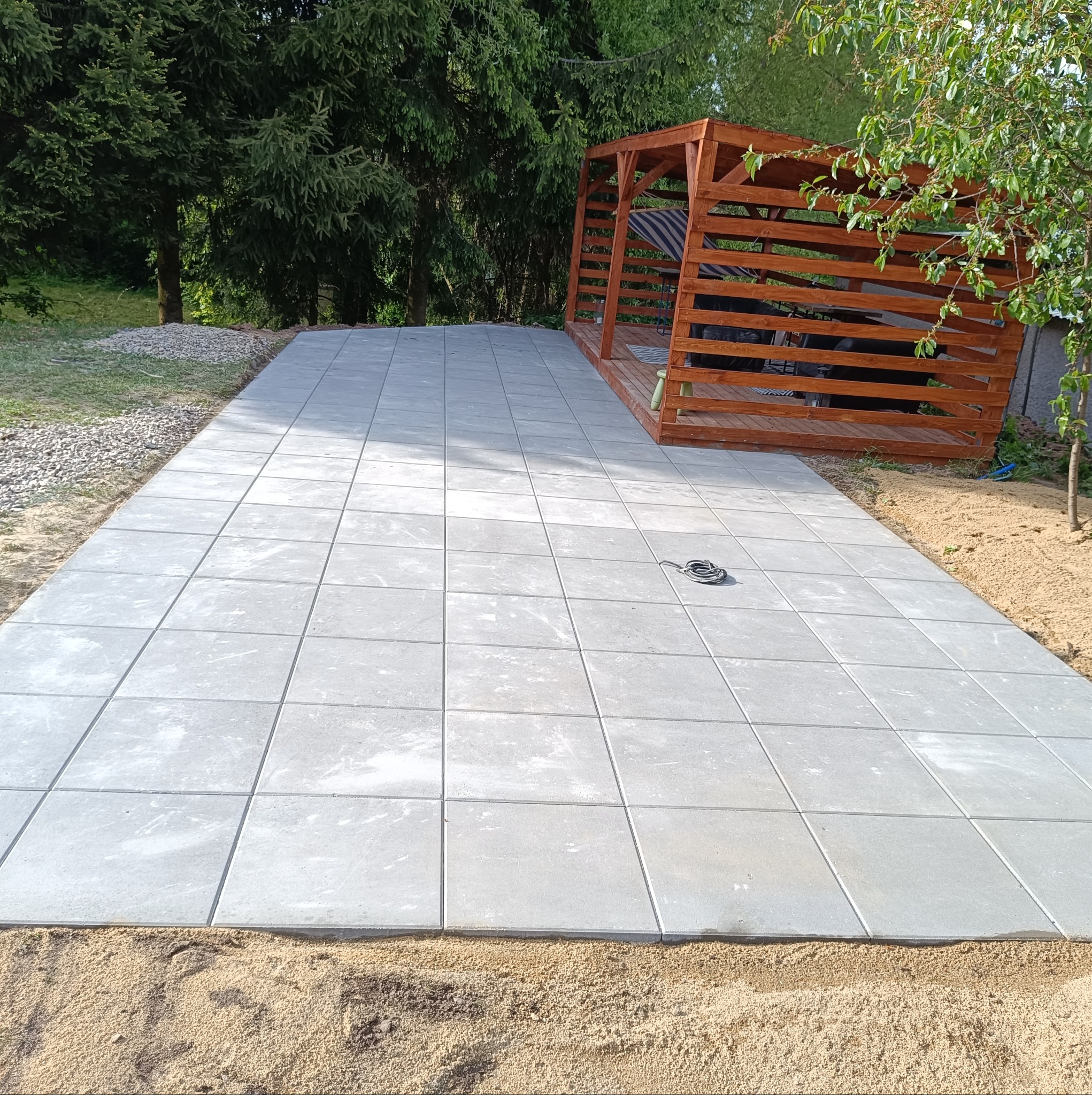 Świeżo ułożona, szara kostka brukowa na posesji z drewnianą altaną w tle. Widoczny piasek wokół kostki i fragment zieleni. Kabel leży na kostce.