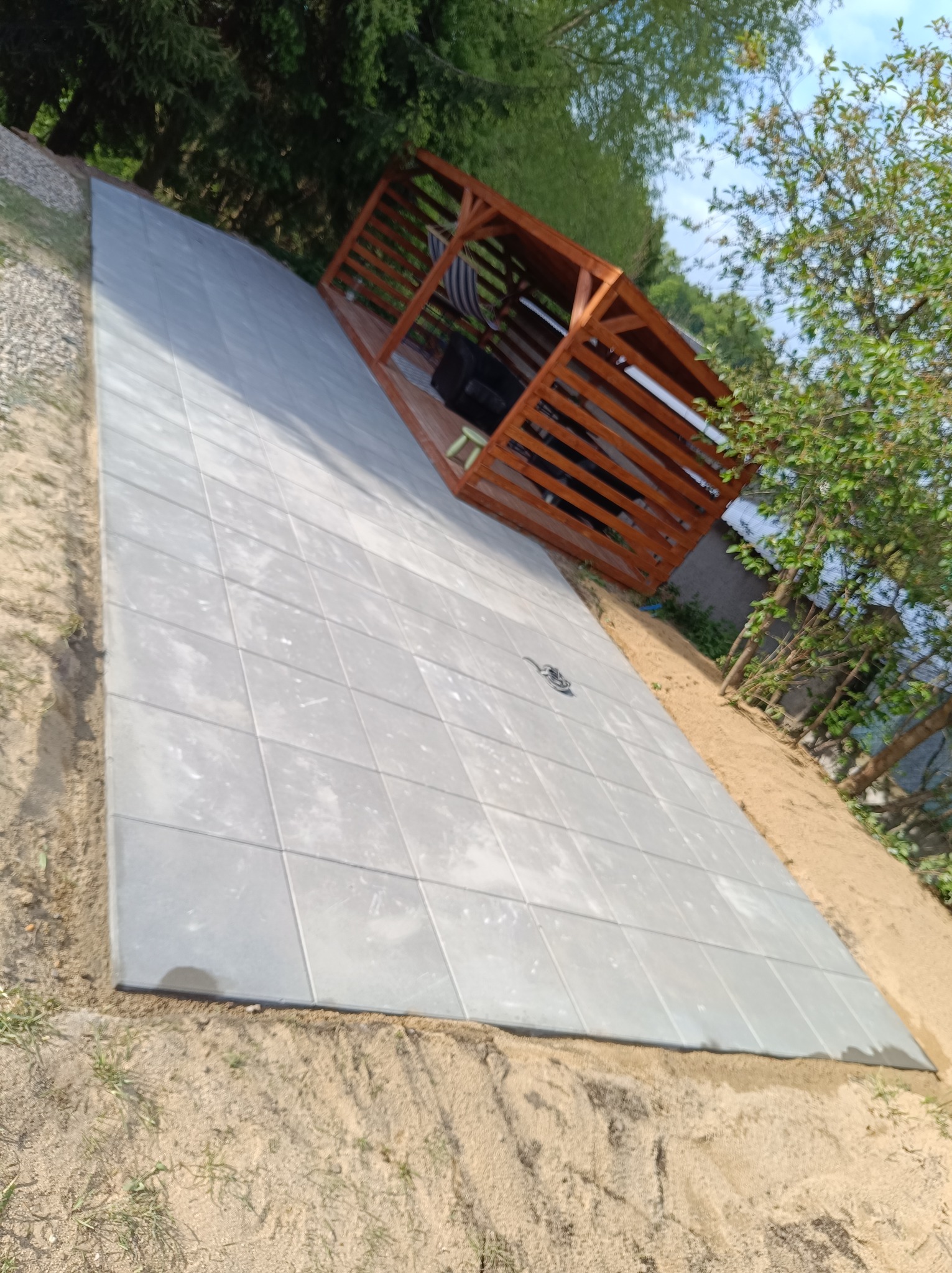 Ułożona, szara kostka brukowa tworząca taras pod drewnianą altaną ogrodową. Widoczne zielone drzewa i piasek.