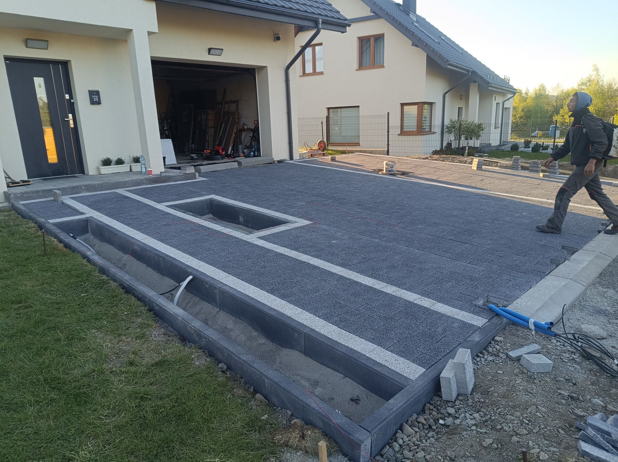 Nowoczesny podjazd z kostki brukowej w trakcie układania, z widocznymi obrzeżami i miejscem na rośliny. W tle dom jednorodzinny i osoba w oddali.