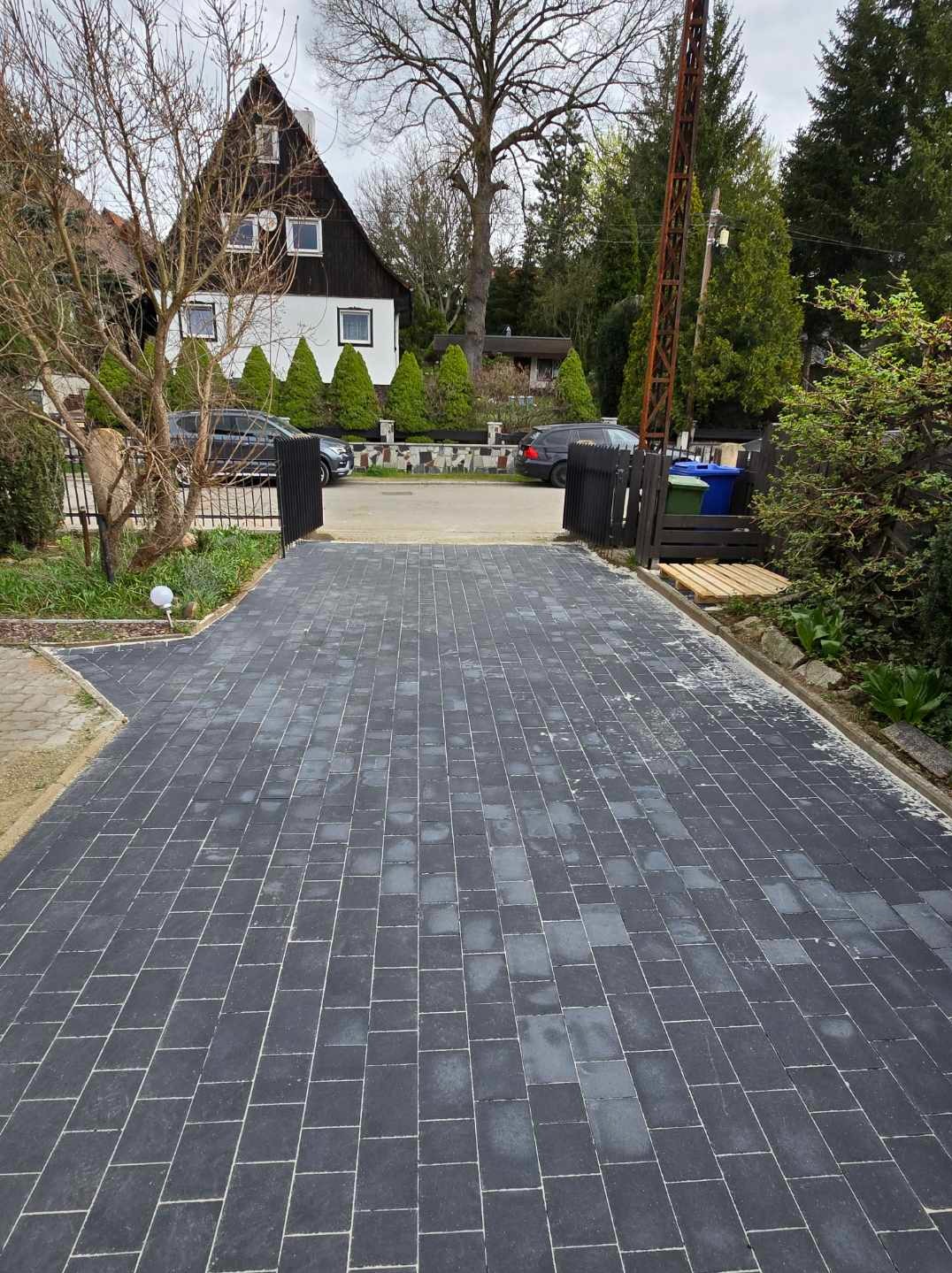 Ułożona, szara kostka brukowa na podjeździe do domu z czarną bramą i zadbaną zielenią. Widok na dom z ciemnymi oknami i drzewami w tle.