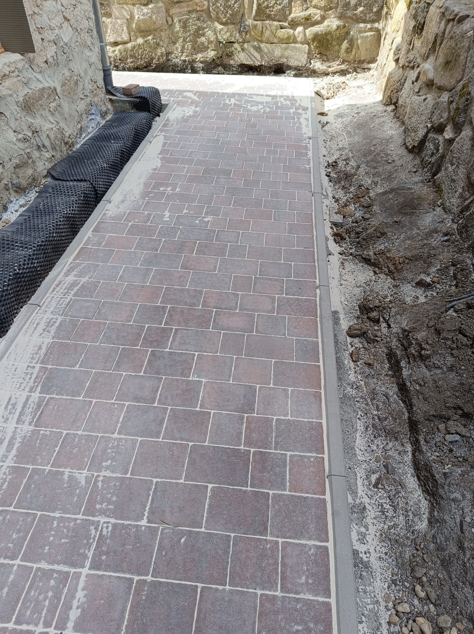 Ułożona kostka brukowa wzdłuż kamiennego muru, z widocznym drenażem i przygotowanym terenem po drugiej stronie. Ciemnobrązowa kostka, szara obrzeża.