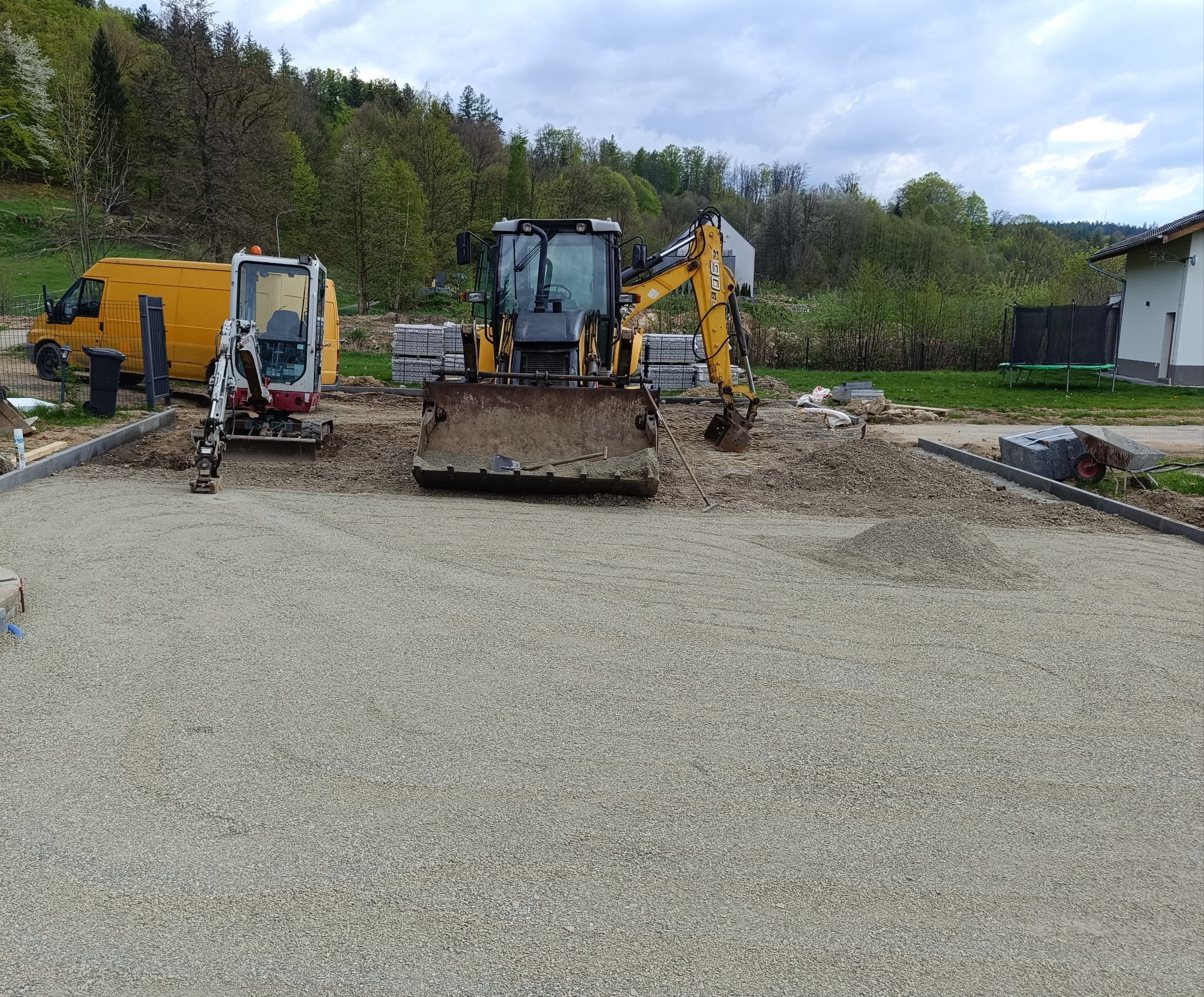 Dwa żółte pojazdy budowlane na żwirowym podłożu, z widocznym stosem kostki brukowej i zielonym tłem drzew. Etap przygotowania podłoża pod kostkę.
