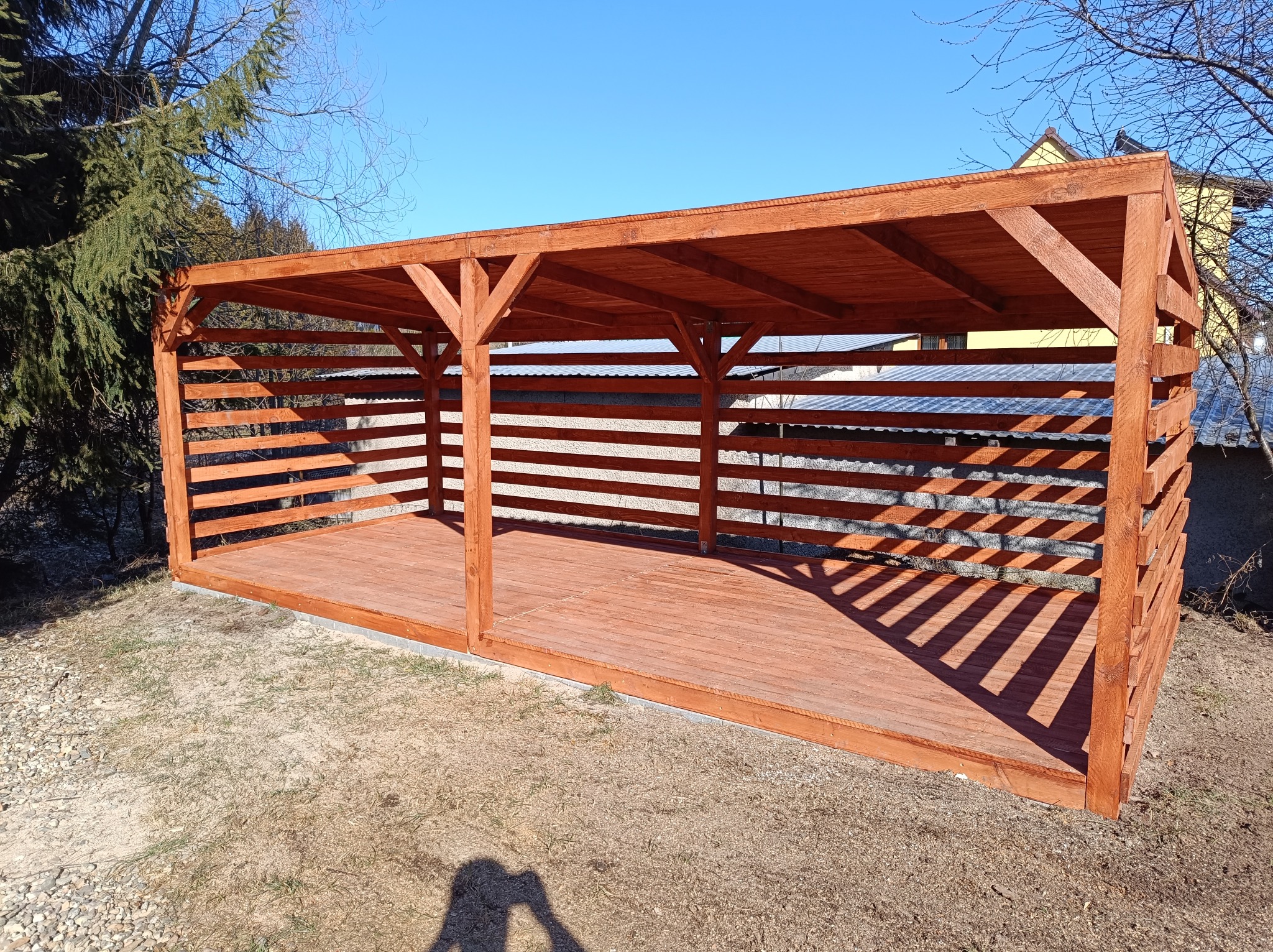 Drewniana, otwarta altana ogrodowa z poziomymi listwami, pomalowana na brązowo, na tle błękitnego nieba. Widoczne drzewa i cień osoby robiącej zdjęcie.