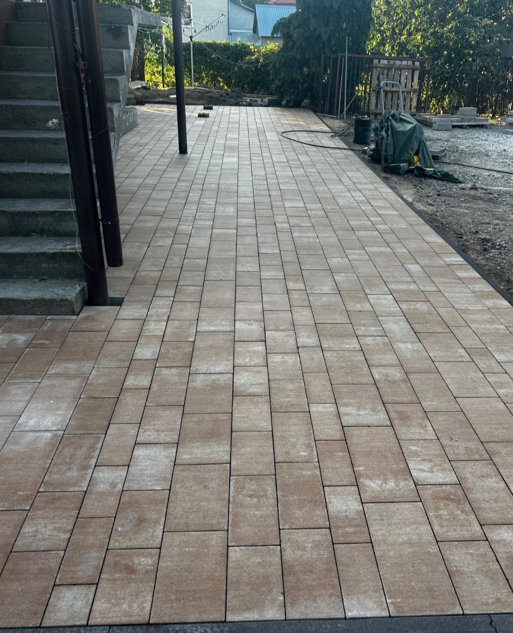 Beżowa kostka brukowa ułożona na posesji, widok z perspektywy schodów. Elementy wyposażenia ogrodowego w tle. Krawężnik oddziela kostkę od terenu.