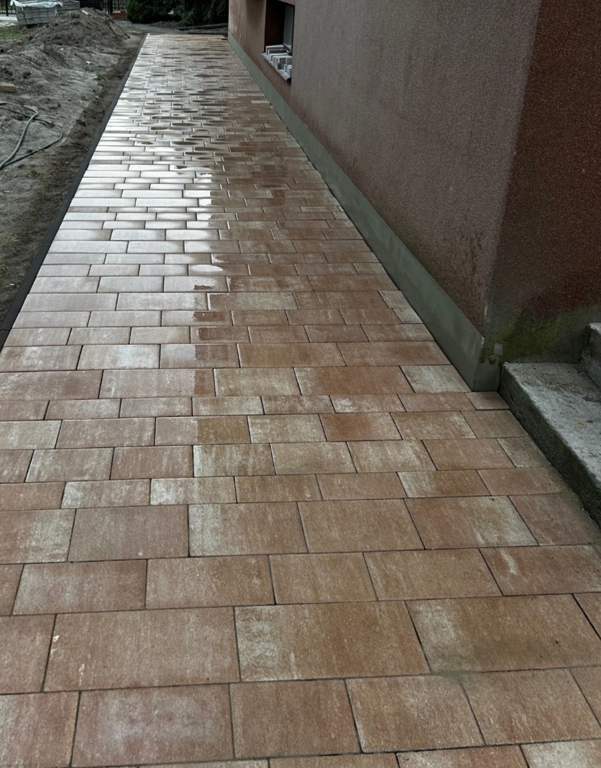 Ułożony chodnik z brązowej kostki brukowej prowadzący wzdłuż budynku z widocznymi schodami i fragmentem terenu przygotowanego pod nasadzenia.