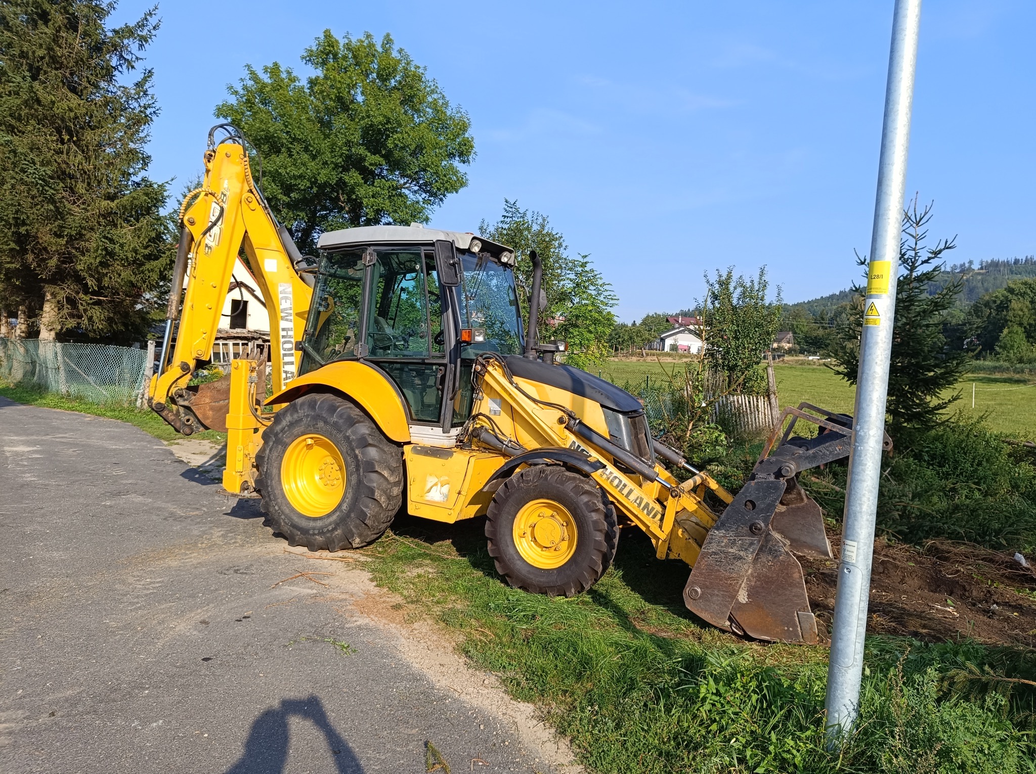 Żółta koparko-ładowarka New Holland zaparkowana na poboczu drogi, z łyżką skierowaną w stronę pryzmy ziemi, na tle zieleni i błękitnego nieba.