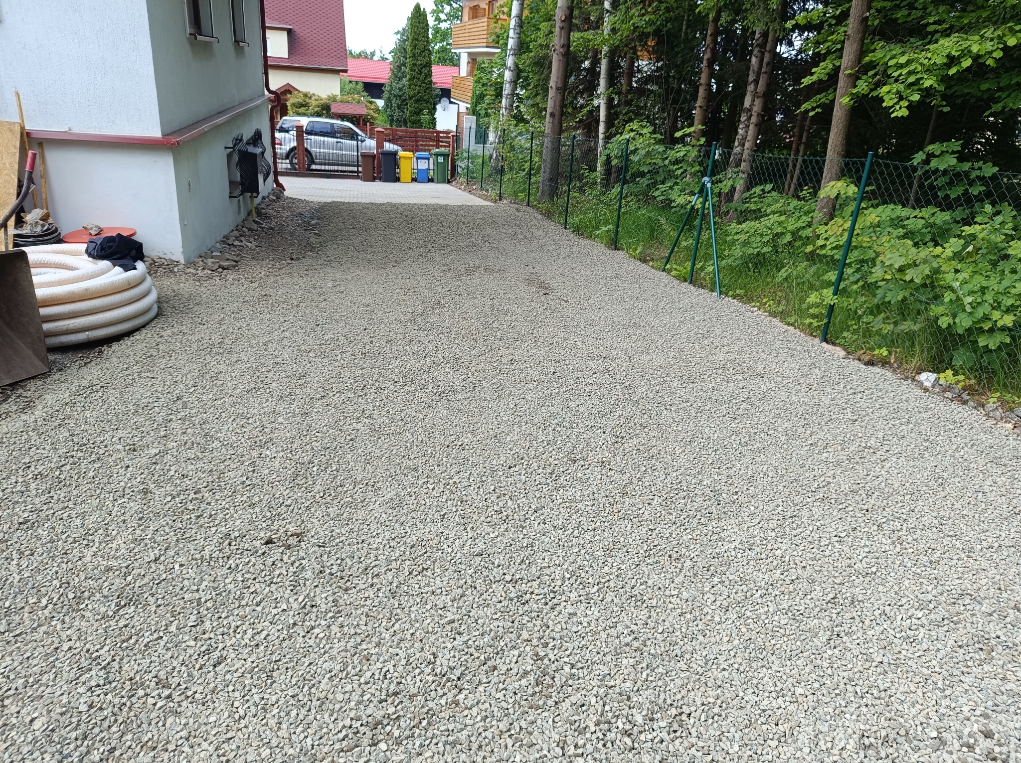Utwardzona żwirem droga dojazdowa, ogrodzona siatką, z widocznymi elementami budynku i drzewami w tle. Na pierwszym planie zwój rur.