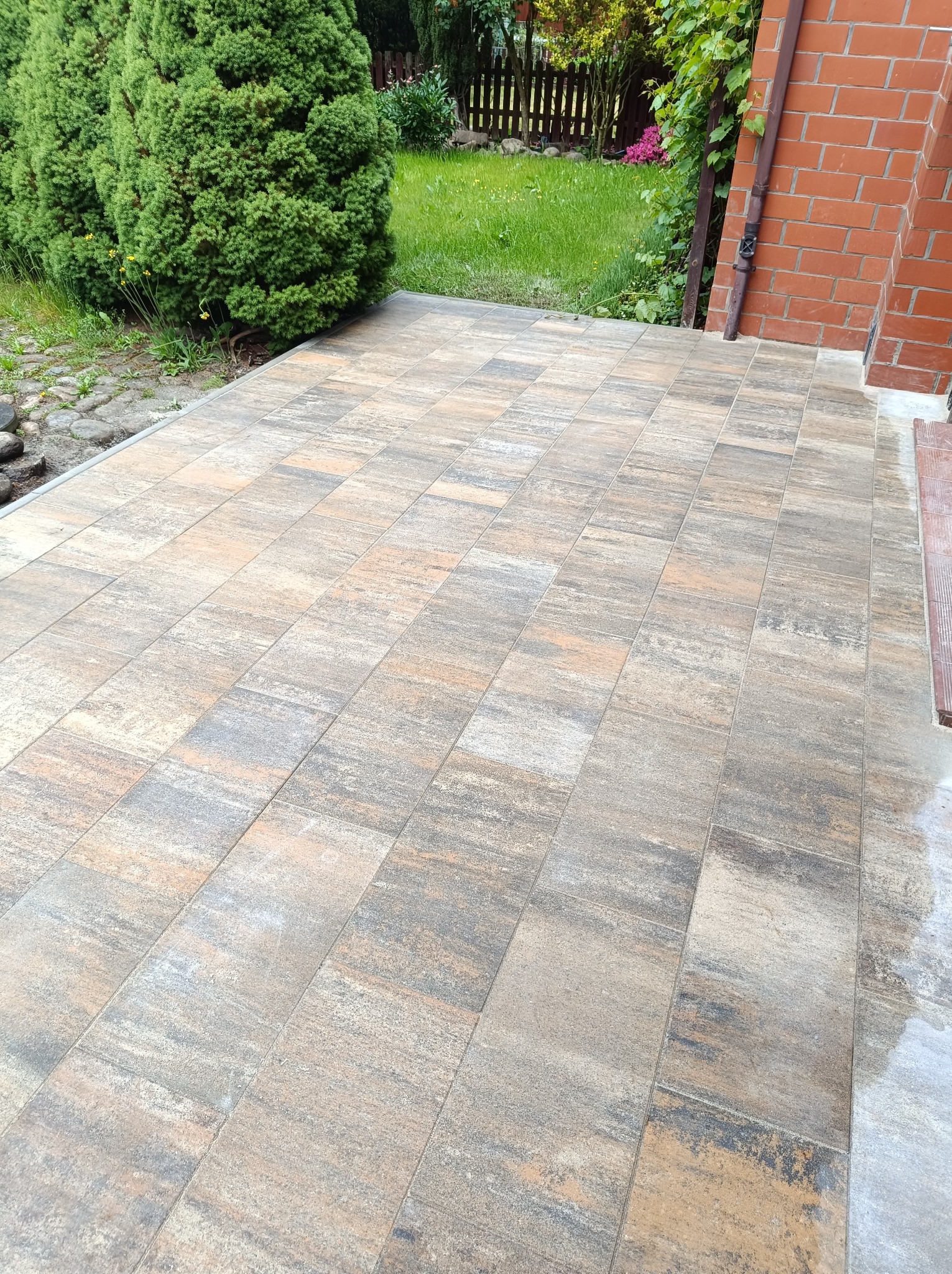 Ułożona kostka brukowa w odcieniach szarości i beżu tworzy estetyczny taras przy budynku z cegły, obok zielony żywopłot i trawnik. Kąt ujęcia podkreśla wzór kostki.