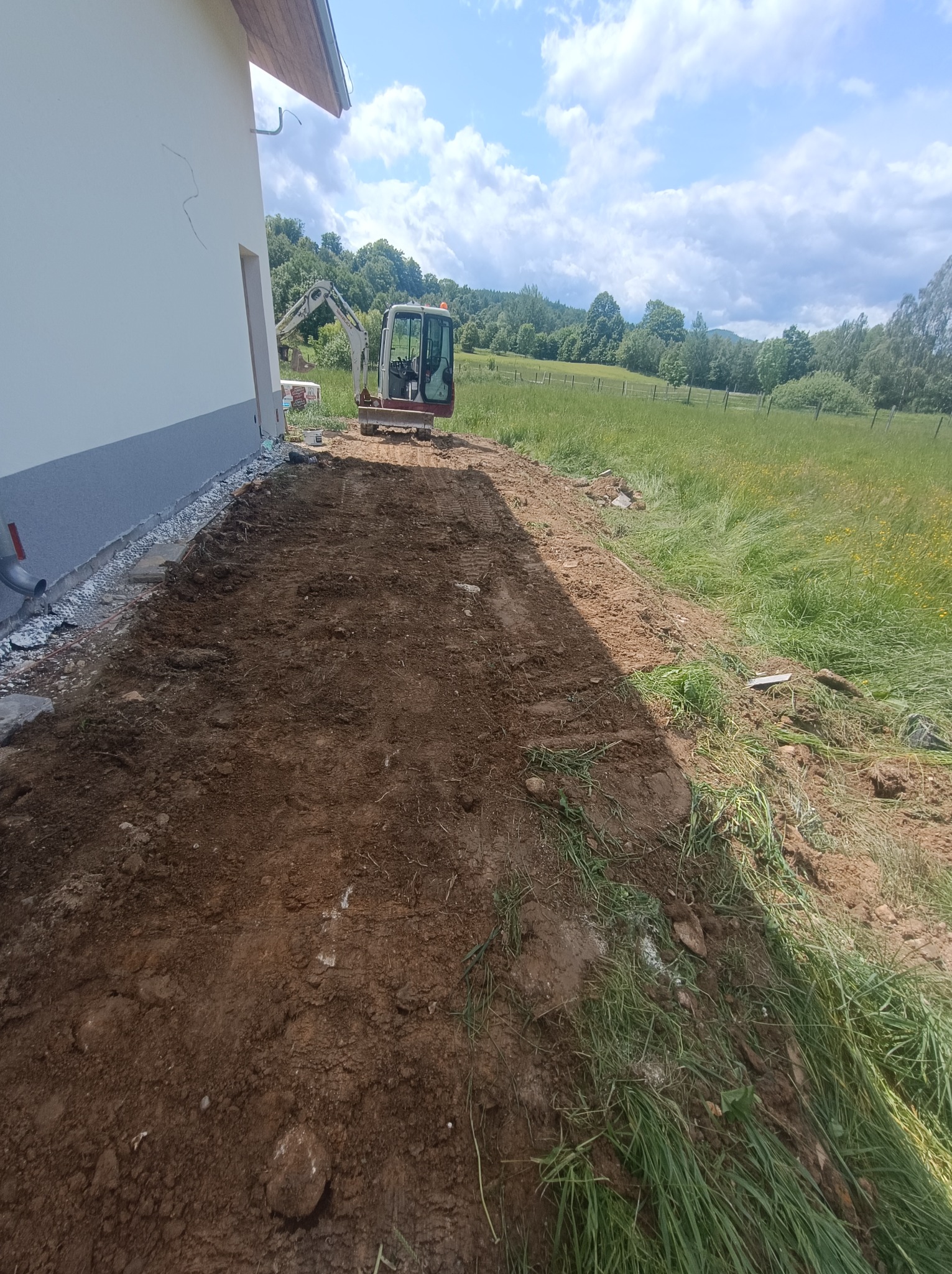 Przygotowany teren pod budowę z widocznym minikoparką i częścią budynku w tle, na tle zieleni. Ślady opon na ziemi.