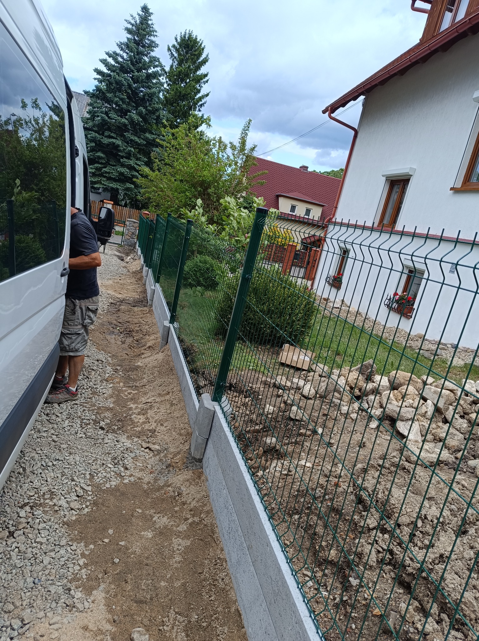 Montaż ogrodzenia panelowego na betonowej podmurówce, widok z boku na ukończony fragment, obok zaparkowany biały bus. W tle dom z czerwonym dachem.