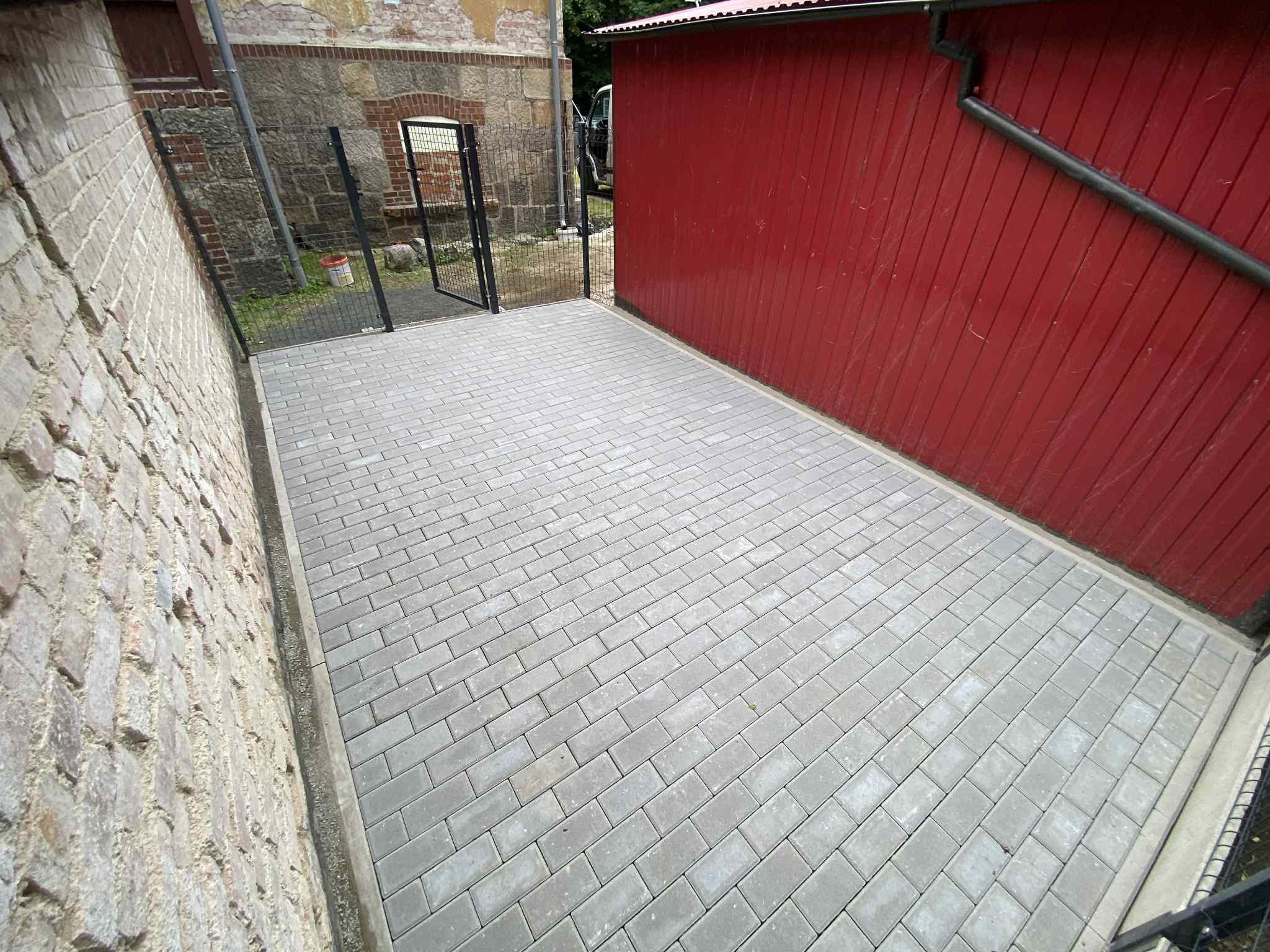 Świeżo ułożona, szara kostka brukowa na podwórku między budynkiem z cegły a czerwonym budynkiem gospodarczym. Widoczna brama z czarnej siatki.