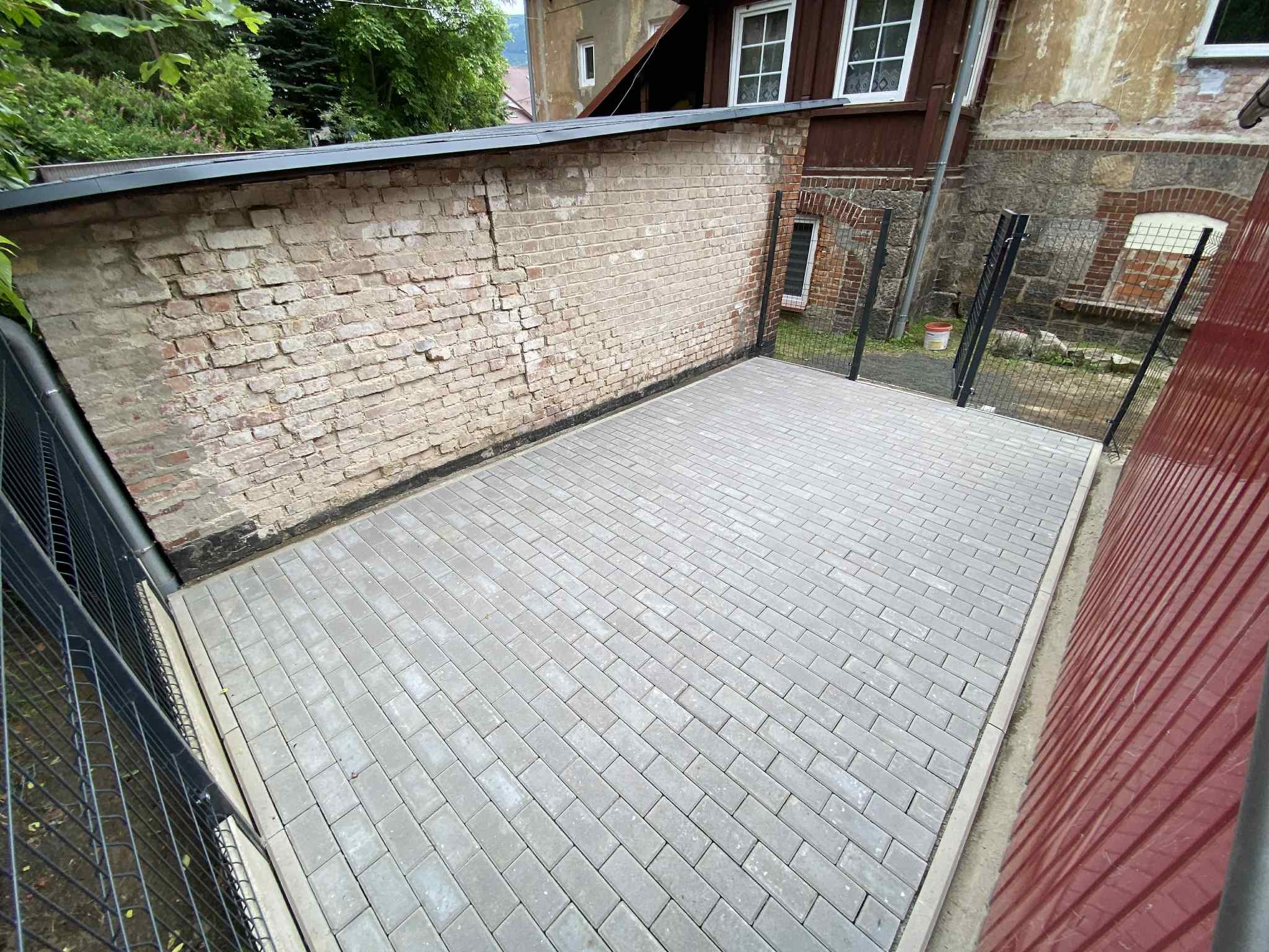 Wyłożony szarą kostką brukową plac między budynkiem z cegły a metalowym ogrodzeniem. Ułożenie w jodełkę. Widoczne detale: fuga, krawężnik.