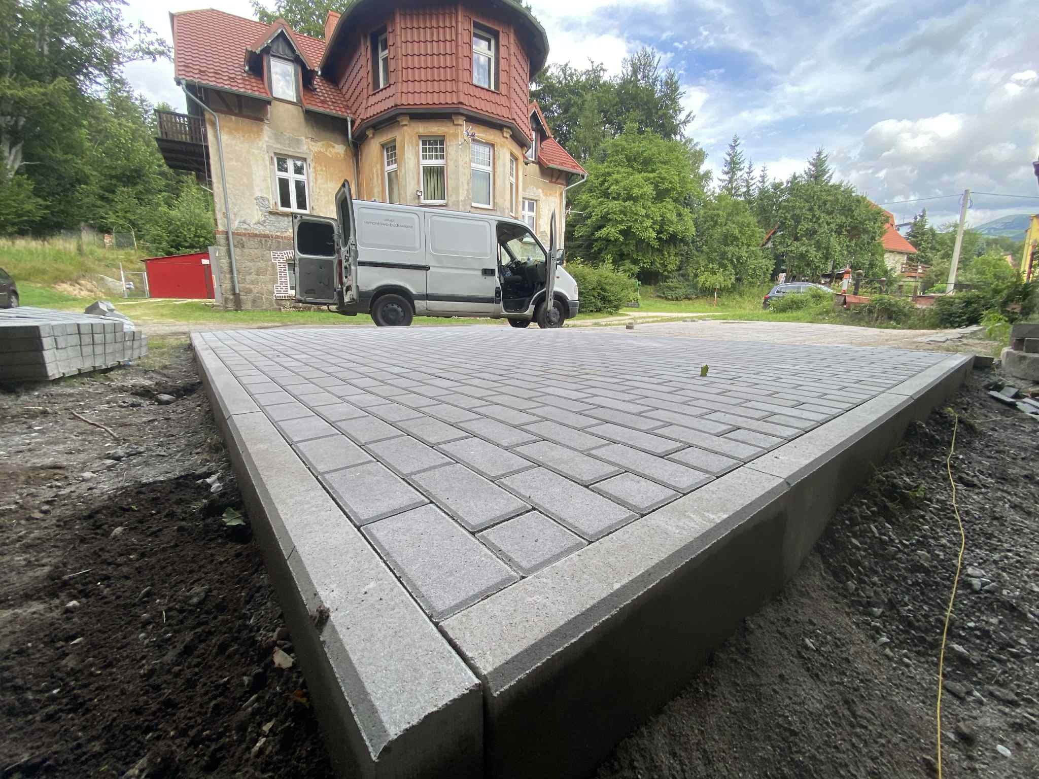 Ułożona szara kostka brukowa z obrzeżami na posesji. W tle budynek z czerwonym dachem i dostawczy samochód z otwartymi drzwiami.