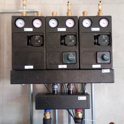 SZEWSKI INSTALATORSTWO - Zamknięty rozdzielacz ogrzewania podłogowego Afriso z manometrami i pompami obiegowymi, zamontowany na ścianie z szarych płytek, widoczne rury doprowadzające.