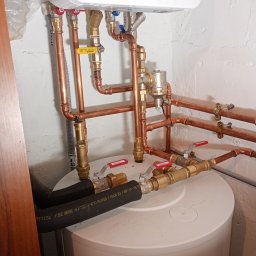 SZEWSKI INSTALATORSTWO - Biały bojler Bosch z miedzianą instalacją hydrauliczną, zawory Calido i czarne węże. Widoczne połączenia i elementy montażowe na tle białej ściany.