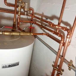 SZEWSKI INSTALATORSTWO - Biały zasobnik ciepłej wody marki Bosch z miedzianą instalacją hydrauliczną, zawory z czerwonymi uchwytami, rury mocowane do ściany. Widok z bliska na instalację.