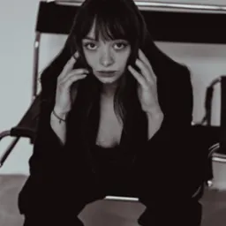 Portret kobiety o długich, ciemnych włosach i piegach, w czarnej marynarce, siedzącej na krześle na tle jasnej ściany. Ujęcie centralne, stonowana kolorystyka.