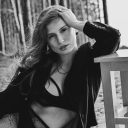 Agnieszka Kenig Fotografia - Czarno-białe, sensualne zdjęcie kobiety w bieliźnie i marynarce, opierającej się o taboret na tle drzew. Widoczny tatuaż na ramieniu, wzrok skierowany w obiektyw.