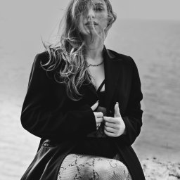 Agnieszka Kenig Fotografia - Czarno-białe zdjęcie kobiety w marynarce i rajstopach w wężowy wzór, z rozwianymi włosami na tle wody, uchwycone w naturalnym oświetleniu.