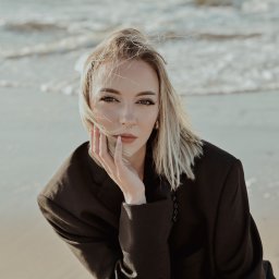 Agnieszka Kenig Fotografia - Portret kobiety w czarnym żakiecie na tle morza. Blond włosy rozwiane, wzrok skierowany w obiektyw, dłoń oparta o policzek. Delikatny makijaż, kolczyk w nosie.