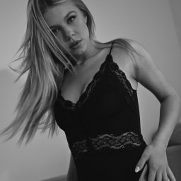 Agnieszka Kenig Fotografia - Czarno-białe ujęcie kobiety w body z koronką, z rozwianymi włosami, spoglądającej w dół, z tatuażem na udzie, uchwycone pod kątem z dołu.