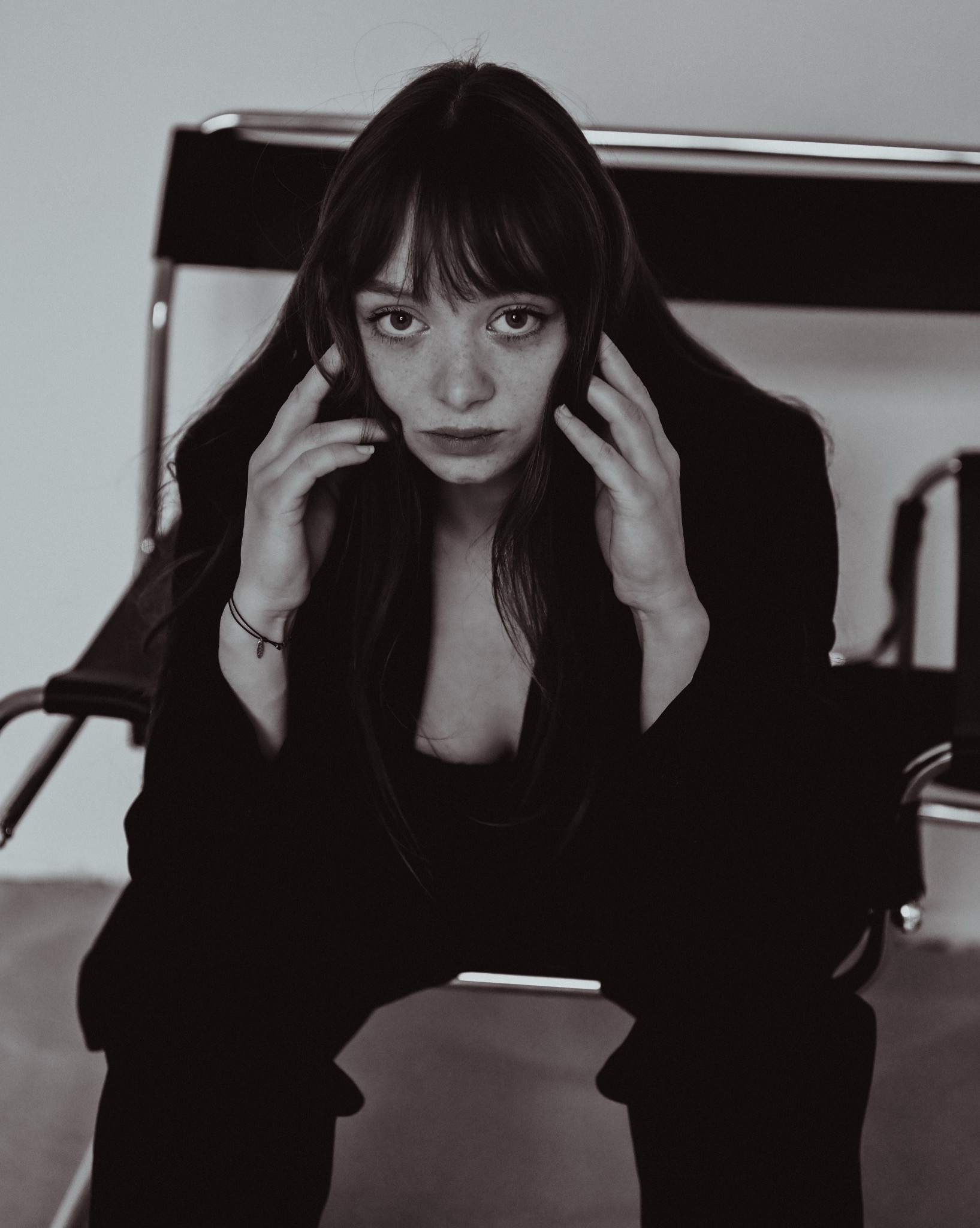 Portret kobiety o długich, ciemnych włosach i piegach, w czarnej marynarce, siedzącej na krześle na tle jasnej ściany. Ujęcie centralne, stonowana kolorystyka.