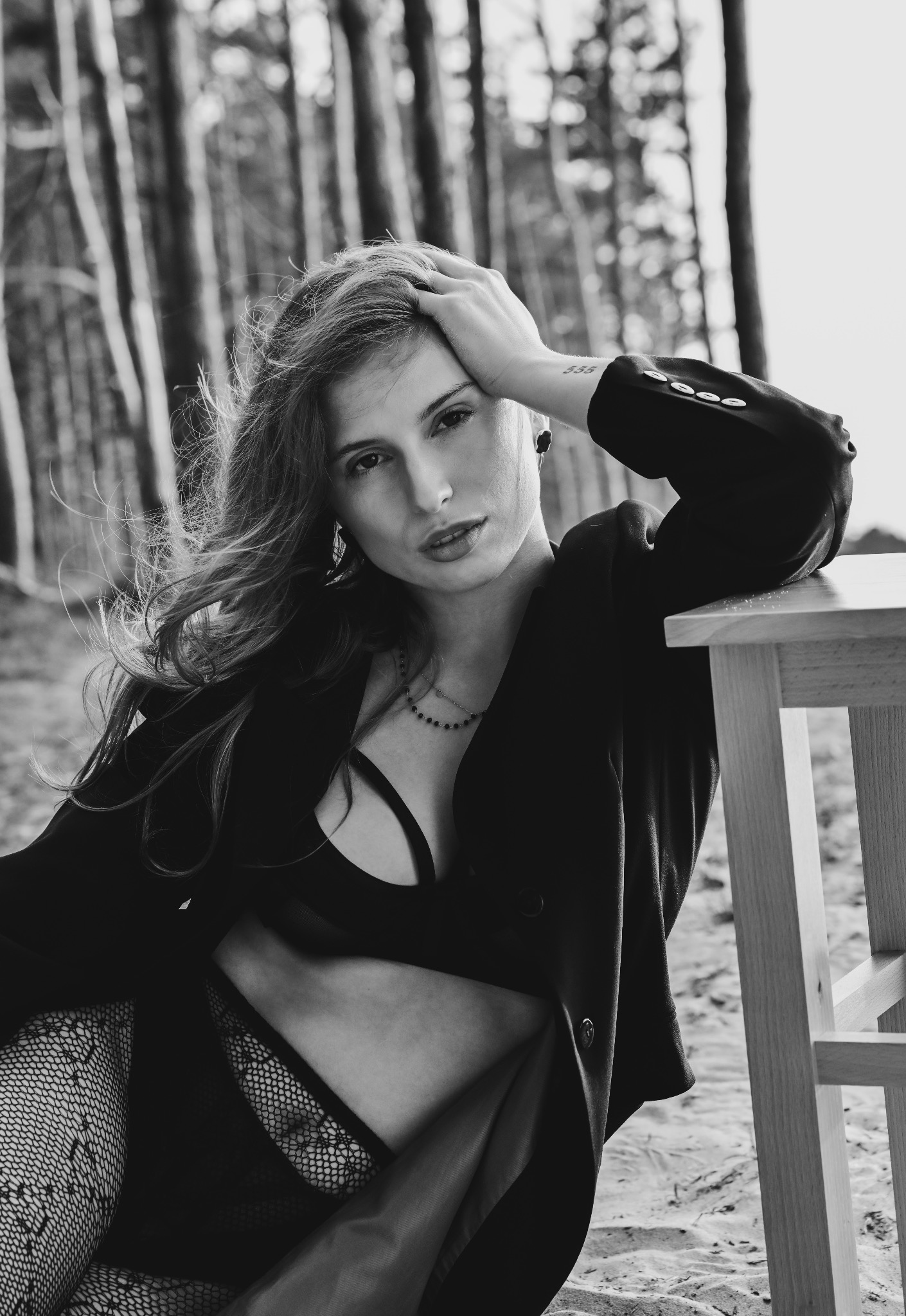 Czarno-białe, sensualne zdjęcie kobiety w bieliźnie i marynarce, opierającej się o taboret na tle drzew. Widoczny tatuaż na ramieniu, wzrok skierowany w obiektyw.