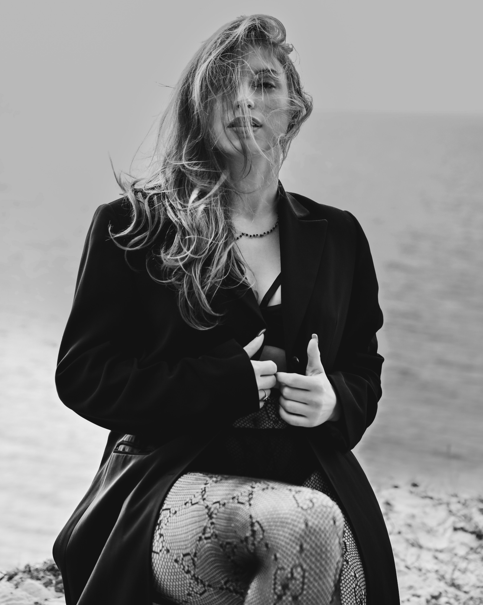 Czarno-białe zdjęcie kobiety w marynarce i rajstopach w wężowy wzór, z rozwianymi włosami na tle wody, uchwycone w naturalnym oświetleniu.