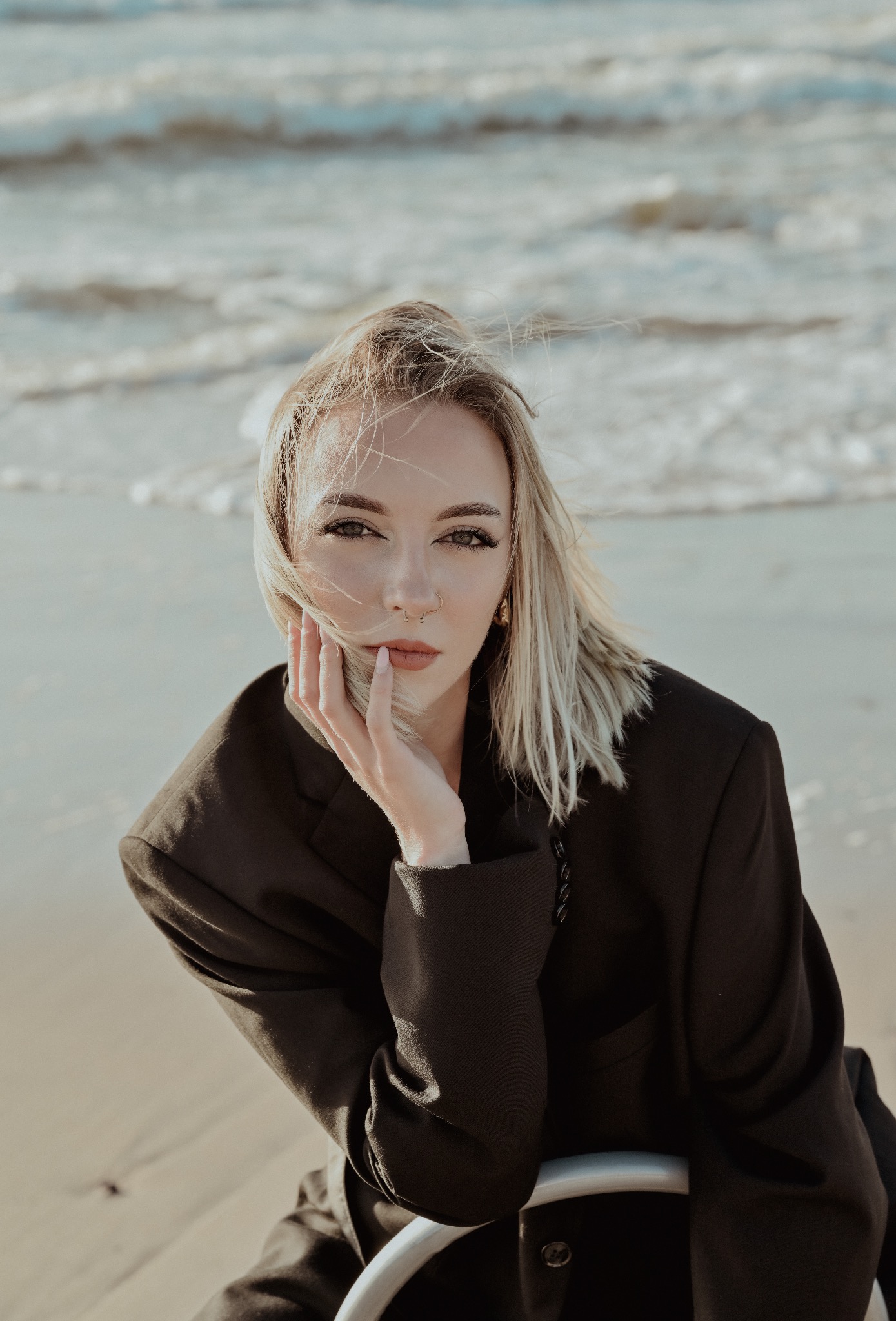 Portret kobiety w czarnym żakiecie na tle morza. Blond włosy rozwiane, wzrok skierowany w obiektyw, dłoń oparta o policzek. Delikatny makijaż, kolczyk w nosie.