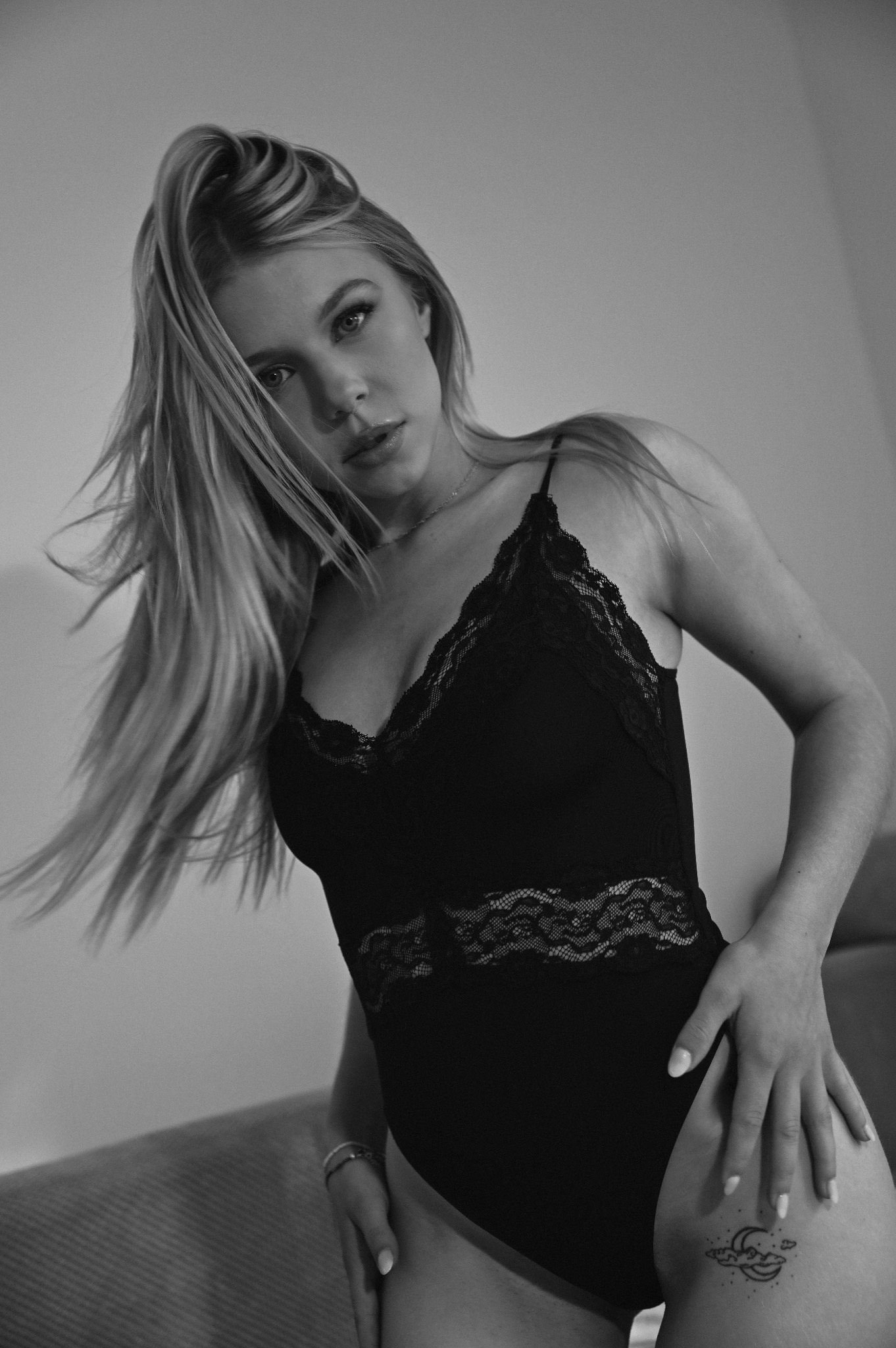 Czarno-białe ujęcie kobiety w body z koronką, z rozwianymi włosami, spoglądającej w dół, z tatuażem na udzie, uchwycone pod kątem z dołu.