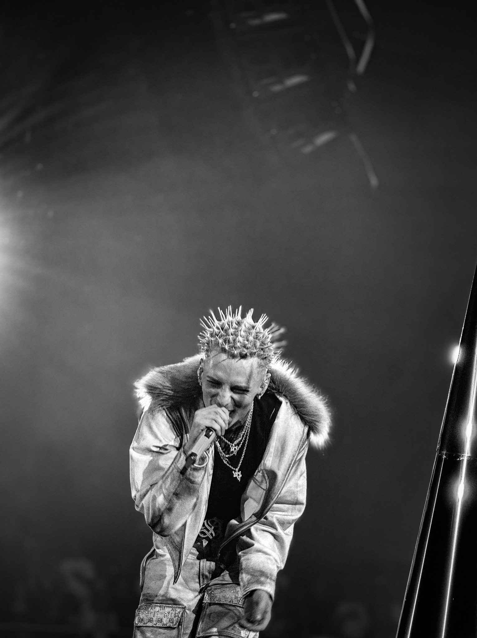 Czarno-białe zdjęcie piosenkarza z kolczastą fryzurą, śpiewającego do mikrofonu na scenie podczas koncertu, w kurtce z futrzanym kołnierzem i łańcuchami na szyi.