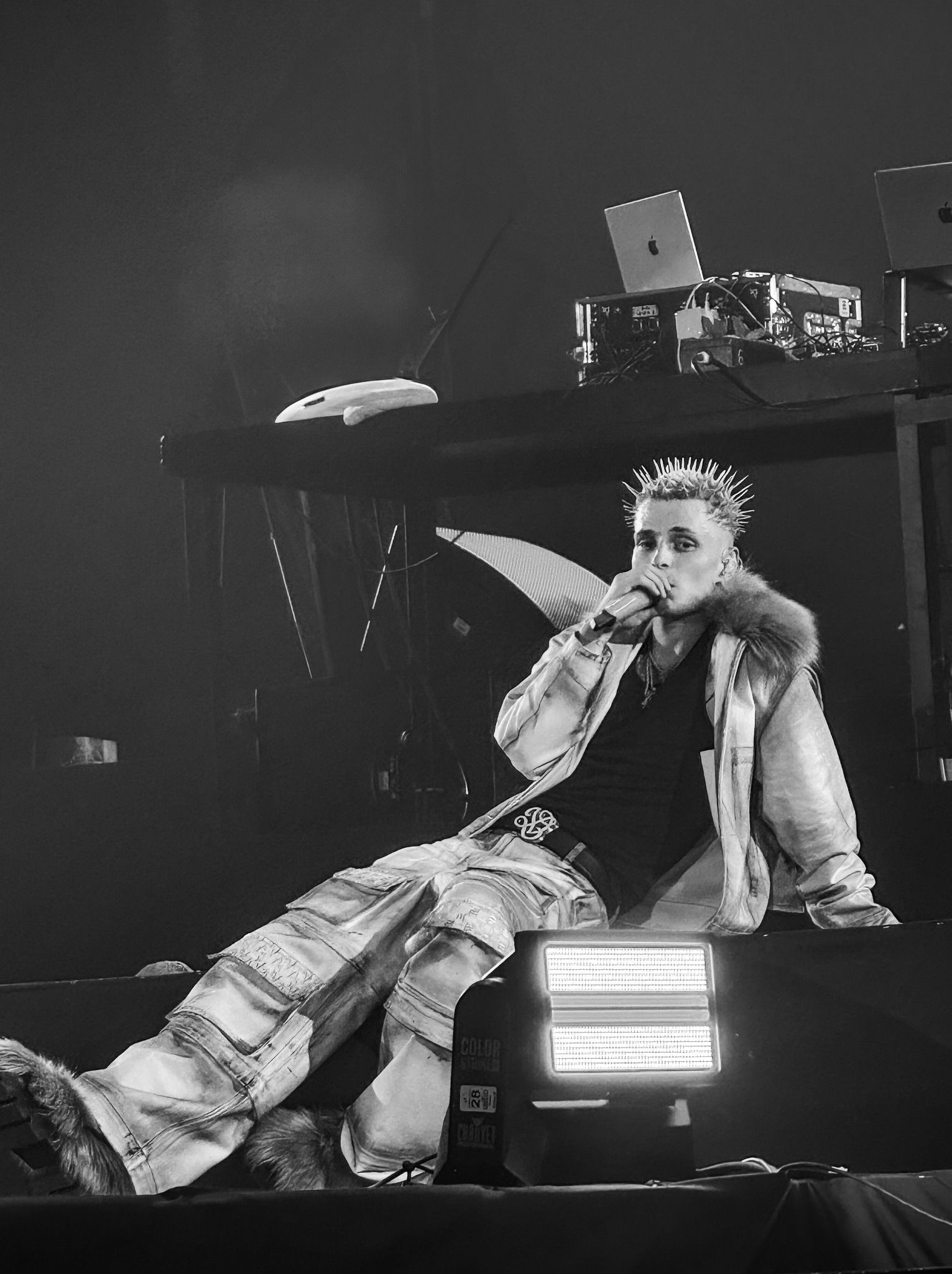 Czarno-białe zdjęcie piosenkarza z kolcami we włosach, siedzącego na scenie z mikrofonem w dłoni, w tle sprzęt muzyczny i oświetlenie sceniczne.