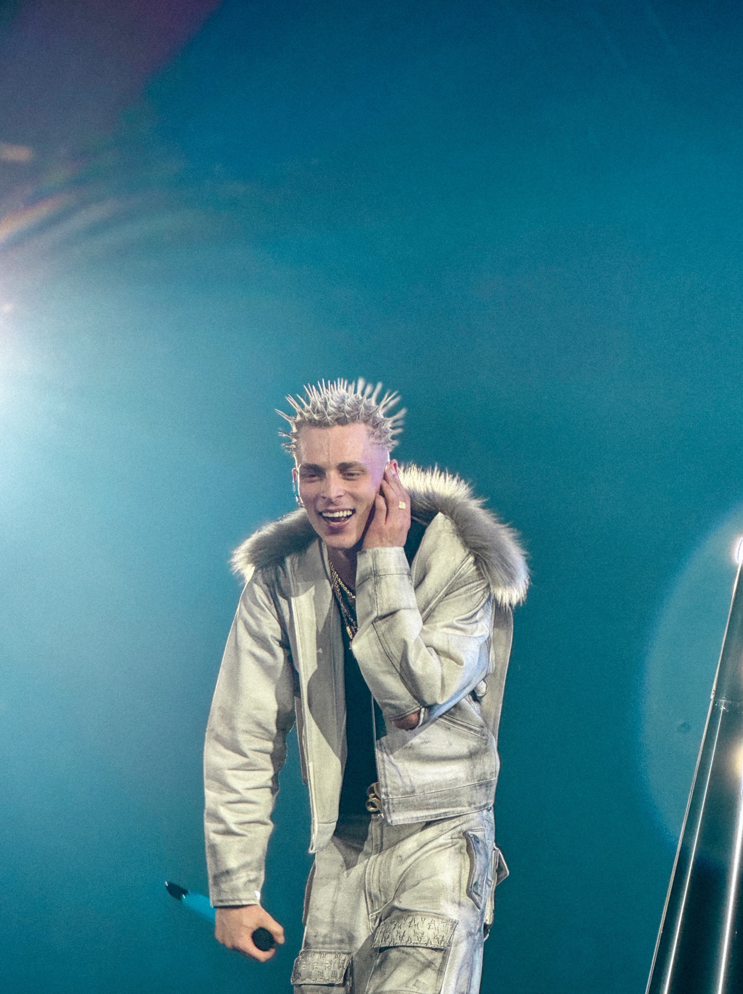 Młody mężczyzna z kolczastymi włosami, w srebrnej kurtce z futerkiem i spodniach cargo, uśmiecha się, trzymając mikrofon na tle turkusowej sceny.