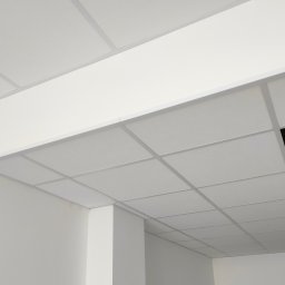 WINBUDPRO - Biały sufit podwieszany kasetonowy z widoczną instalacją wentylacyjną w otworze, w jasnym pomieszczeniu biurowym. Minimalistyczny design, równe linie.