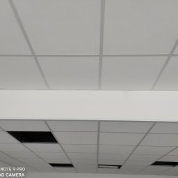 WINBUDPRO - Biały sufit podwieszany z widocznymi panelami i czarnymi otworami wentylacyjnymi. Ujęcie z dołu, perspektywa zbieżna. Oświetlenie równomierne.