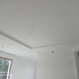 WINBUDPRO - Białe wnętrze po wykończeniu ścian i sufitu, widoczne wystające kable elektryczne, okno i otwór drzwiowy. Minimalistyczny design, surowe wykończenie.