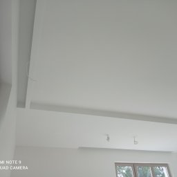 WINBUDPRO - Nowoczesny, biały sufit podwieszany z oświetleniem w trakcie instalacji. Widoczne kable, okno i minimalistyczny design.