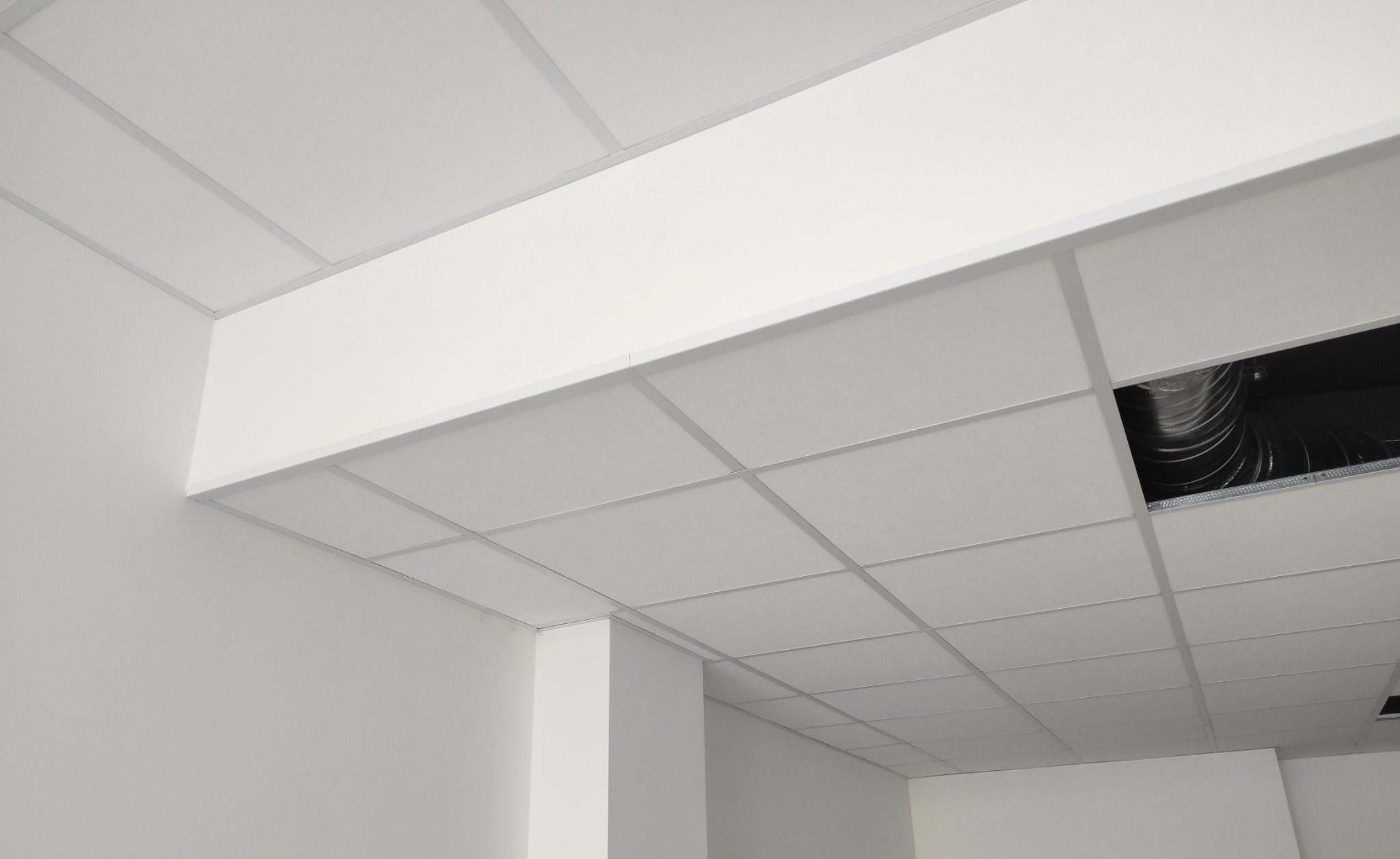 Biały sufit podwieszany kasetonowy z widoczną instalacją wentylacyjną w otworze, w jasnym pomieszczeniu biurowym. Minimalistyczny design, równe linie.