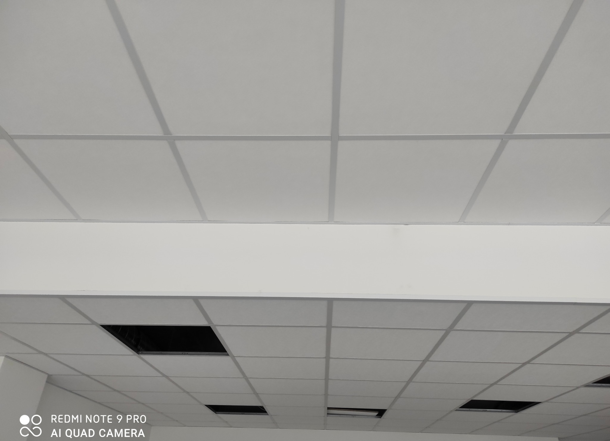 Biały sufit podwieszany z widocznymi panelami i czarnymi otworami wentylacyjnymi. Ujęcie z dołu, perspektywa zbieżna. Oświetlenie równomierne.