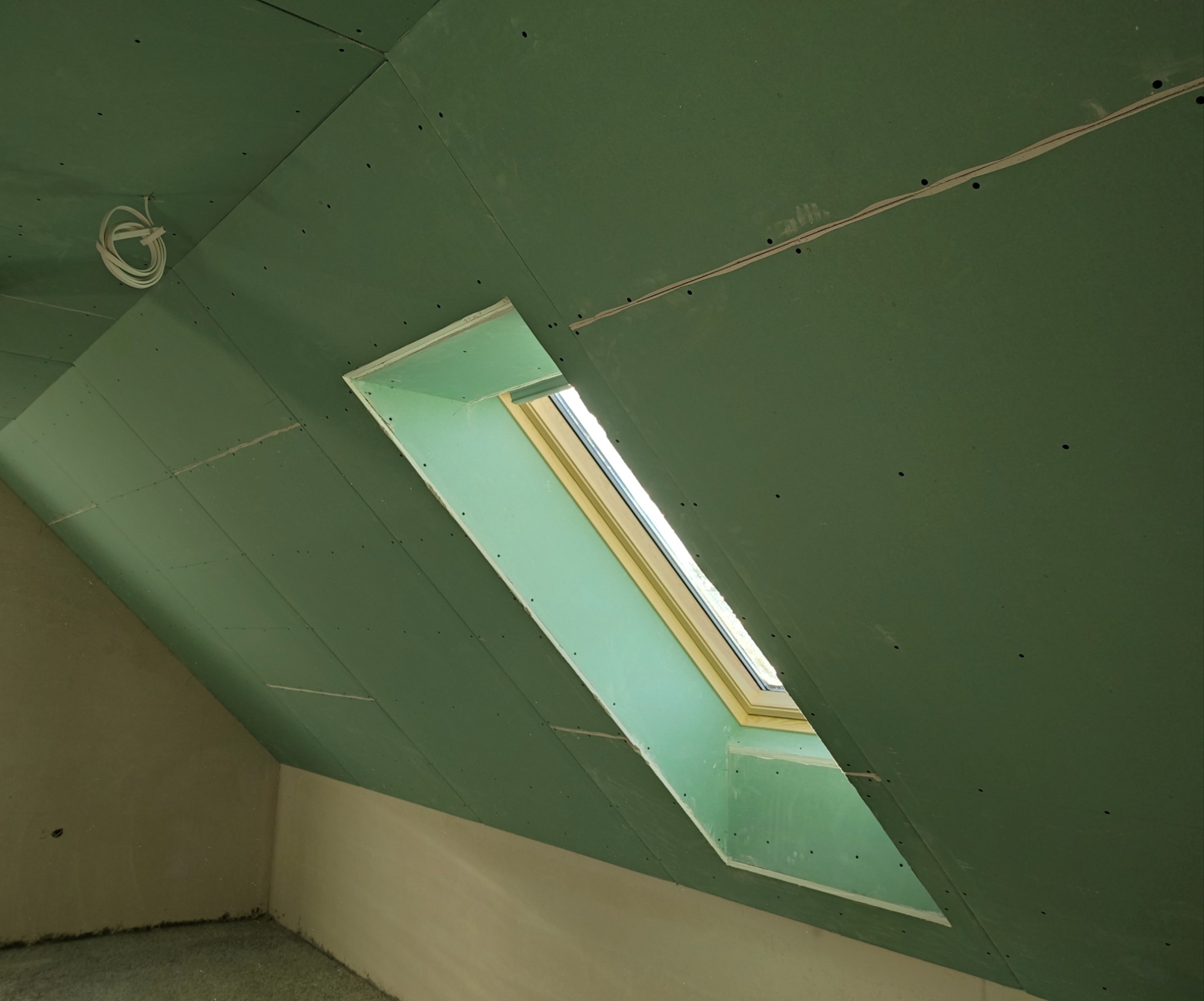 Zielone płyty G-K na poddaszu ze skośnym oknem dachowym. Widoczne łączenia płyt i wystające przewody. Surowy stan wykończenia.