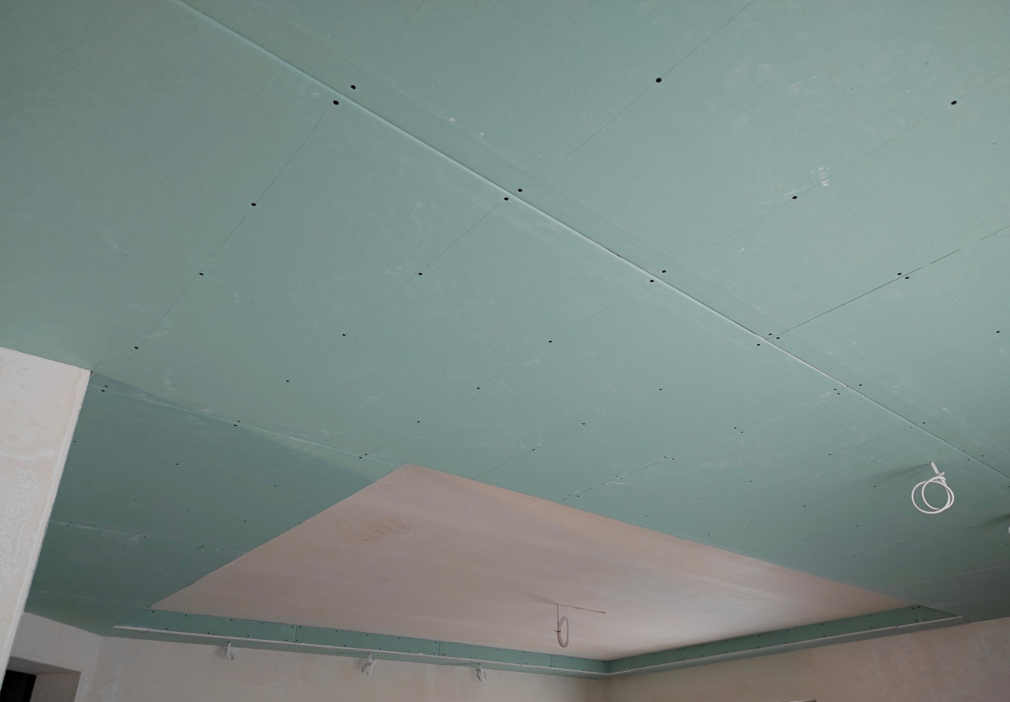 Zielona płyta gipsowo-kartonowa zamontowana na suficie. Część sufitu z wykończoną, gładką powierzchnią. Widoczne łączenia płyt i otwory po wkrętach.