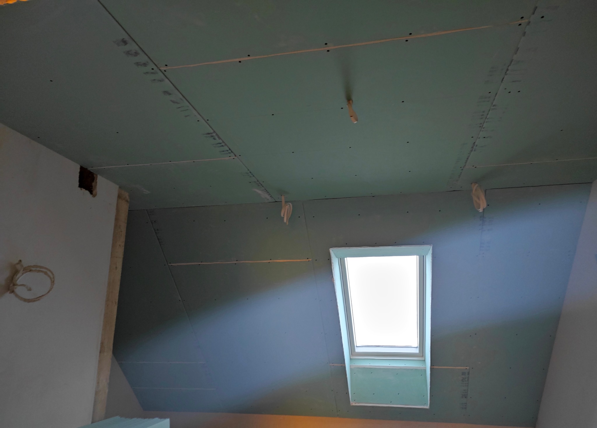 Poddasze z oknem dachowym w trakcie obkładania płytami gipsowo-kartonowymi. Widoczne łączenia płyt i otwory po wkrętach. Fragment instalacji elektrycznej.