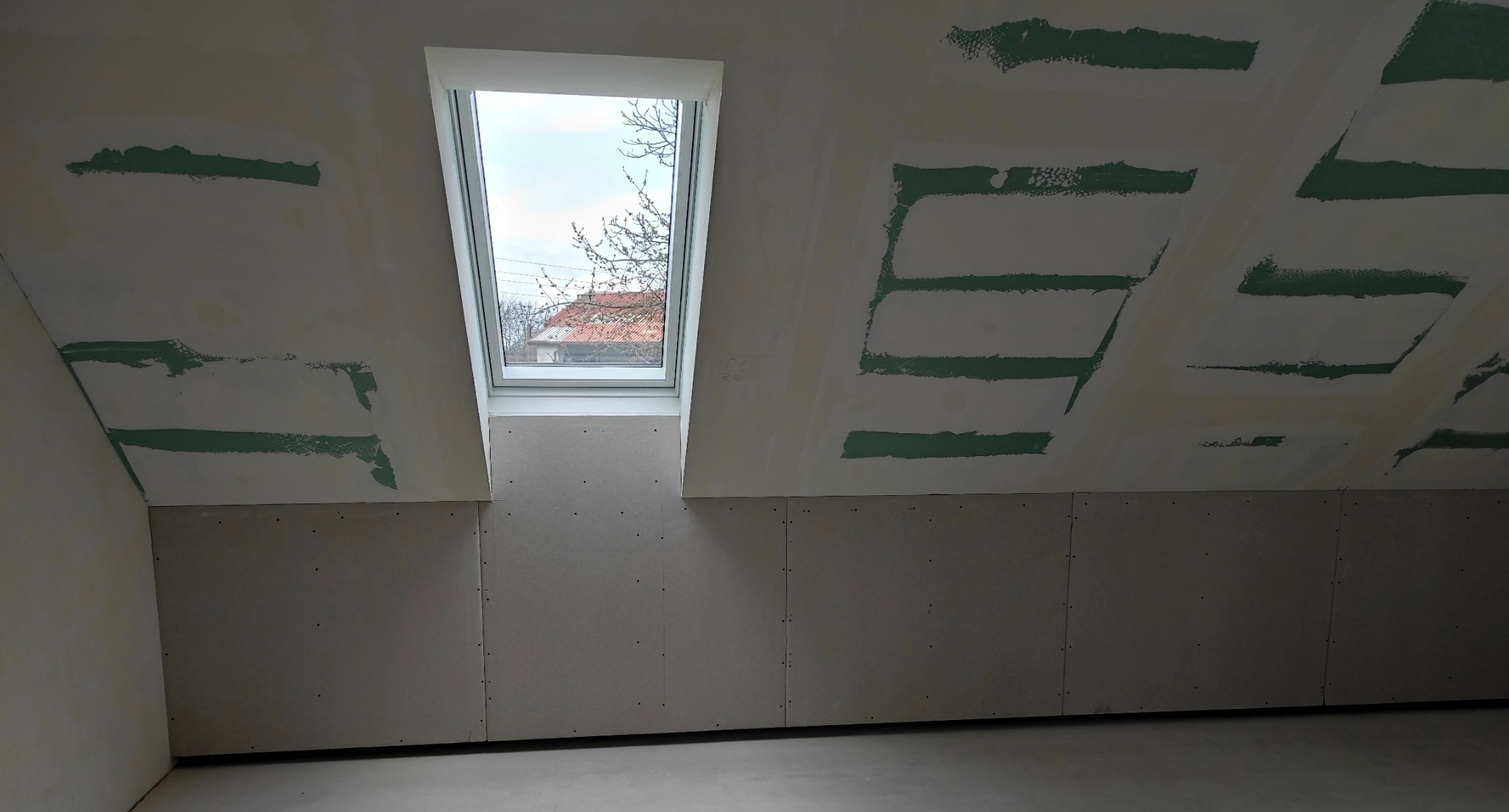 Poddasze z oknem dachowym w trakcie wykańczania płytami karton-gips, widoczne szpachlowane łączenia na skośnych ścianach i suficie. Surowy stan, przygotowanie do malowania.