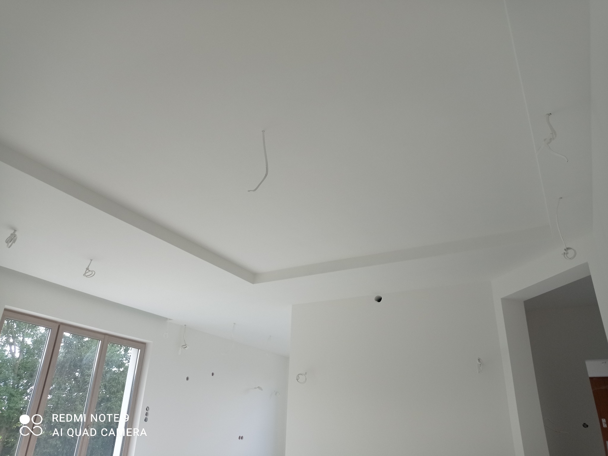 Białe wnętrze po wykończeniu ścian i sufitu, widoczne wystające kable elektryczne, okno i otwór drzwiowy. Minimalistyczny design, surowe wykończenie.