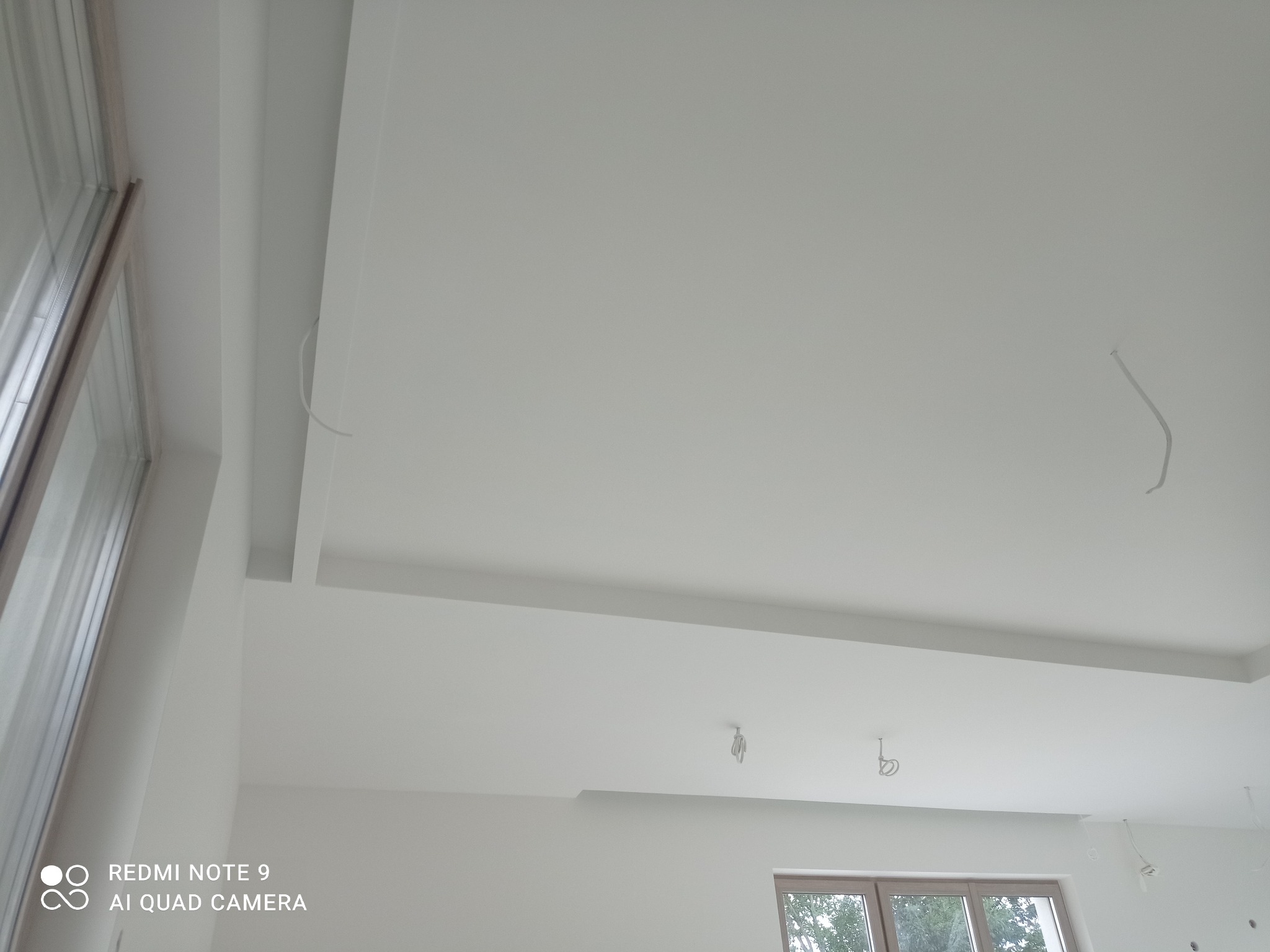 Nowoczesny, biały sufit podwieszany z oświetleniem w trakcie instalacji. Widoczne kable, okno i minimalistyczny design.